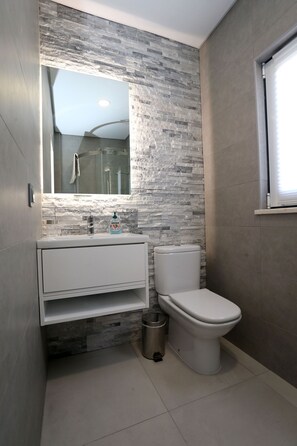 A modern bathroom with a stone wall backsplash, a white sink, a toilet, and a trash can.