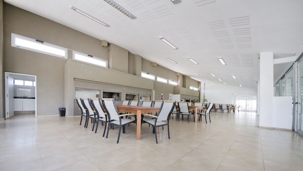 A spacious indoor area with multiple tables and chairs, high ceilings, and large windows allowing natural light. - Milénica Flat 8PJ (Malagueño)