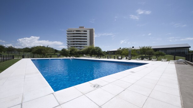 A rectangular swimming pool with white tiled edges, surrounded by a paved area with lounge chairs and a building in the background. - Milénica Flat 4PJ (Malagueño)