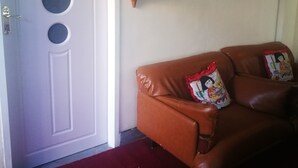 A room with a white door featuring circular cutouts, a brown leather sofa with decorative pillows, and a wall-mounted basket. - Naisar Royal White Apartment (Germiston)