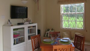 Een eetruimte met een houten tafel, stoelen, een televisie op een wit kastje, en een raam met uitzicht op groen.