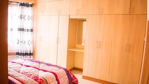 A bedroom with a bed, wooden wardrobe, and a small shelf. - Amani homestay (Syokimau)