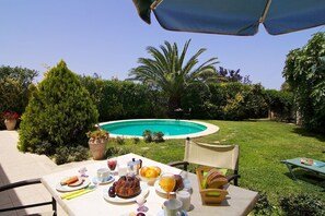 A poolside dining area with a table set for breakfast, surrounded by lush greenery and a clear blue pool. - Stone villa with pool & fantastic sea views (with special autumn offers) (Rethymnon)