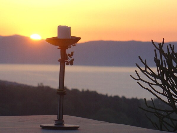 A candle holder with a lit candle on a table, overlooking a body of water and mountains during sunset.