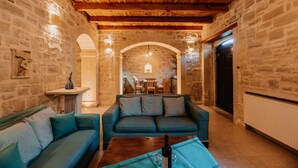 A cozy living room with stone walls, wooden ceiling beams, a sofa, a coffee table with a bottle, and a fireplace. - Stone villa with pool & fantastic sea views (with special autumn offers) (Rethymnon)