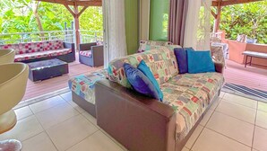 A cozy living area with a sofa, a bench, and a table. There are curtains on the windows, and the room has a tiled floor.