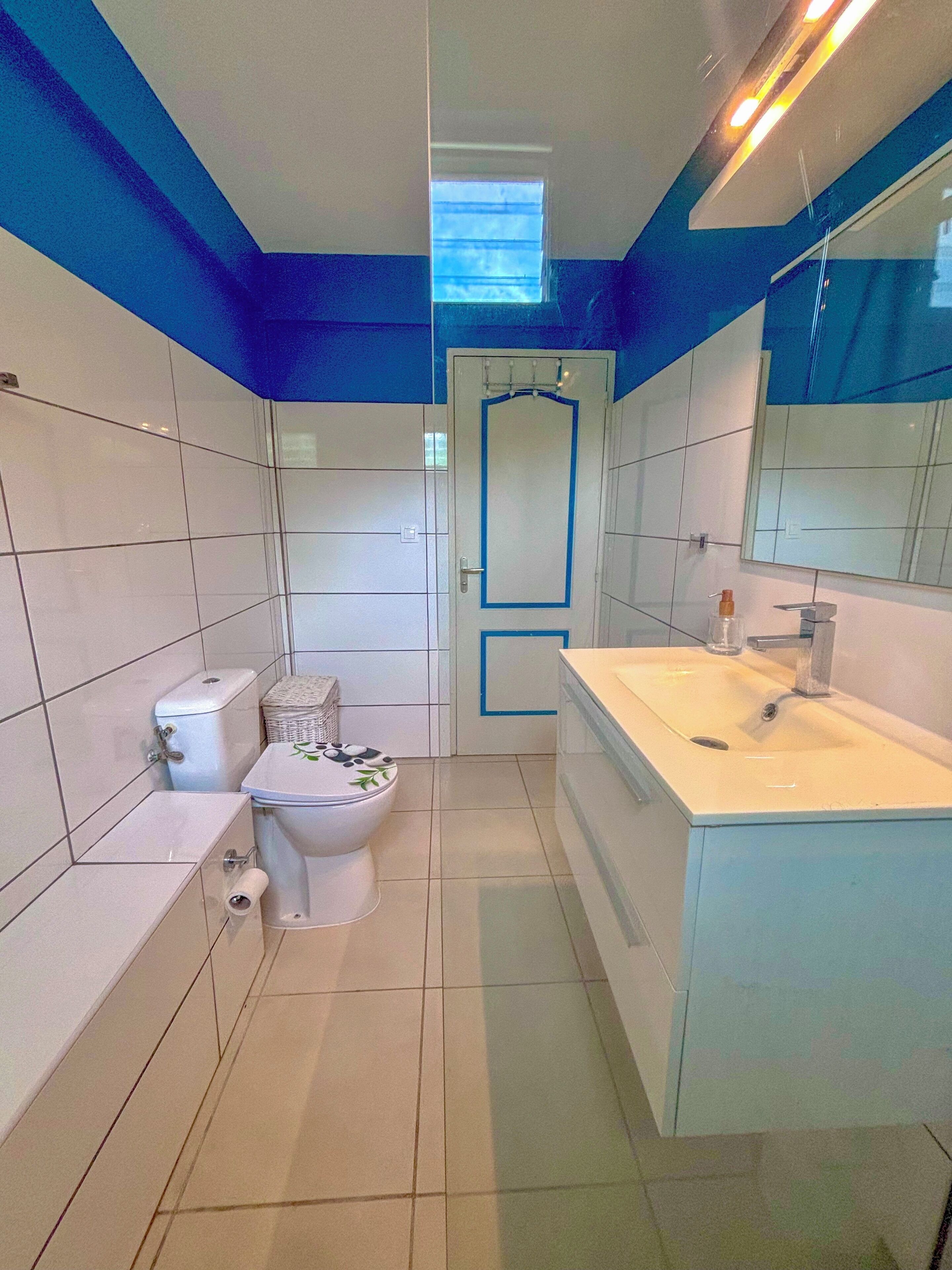 Een badkamer met witte en blauwe tegels, een toilet, een wastafel met spiegel en een raam boven het toilet.