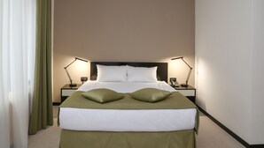 Quarto de hotel com uma cama grande, duas mesas de cabeceira com luminárias e cortinas verdes.