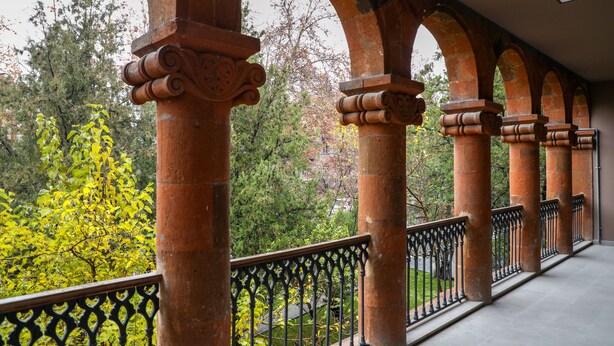 Varanda externa com grades de ferro forjado, sustentada por colunas de pedra vermelha com capitéis ornamentados.