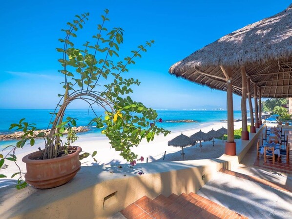 A beachside restaurant with a thatched roof, outdoor seating, and a view of the ocean.