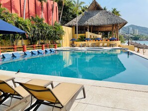 A pool with lounge chairs, a thatched-roof structure, and a view of the sea.