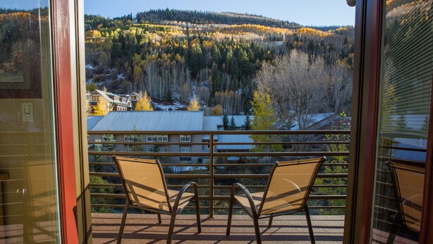 A balcony with two chairs and a view of a forested hillside and buildings.