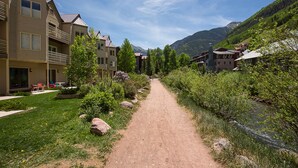 A dirt path leading through a residential area with houses on either side, a river on the right, and mountains in the background.