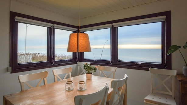 Un coin repas avec une table en bois et des chaises blanches, de grandes fenêtres donnant sur la mer et une lampe suspendue.