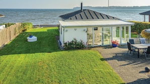 Une véranda avec un toit en verre donnant sur un plan d’eau, une pelouse et un espace patio aménagé avec du mobilier d’extérieur.