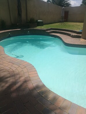 A swimming pool with a curved edge, surrounded by a brick patio. - Naisar Sparkling White Apartment (Germiston)
