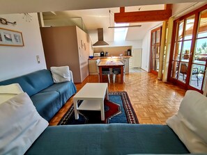 A cozy living room with a blue sofa, a small white table, and a kitchen area with a dining table and chairs. - Landhaus Mitsche, apartment panoramic view in a quiet, sunny position (Radnig)