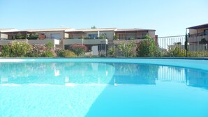 A swimming pool with a clear blue water surface, surrounded by a metal fence. In the background, there are multi-story buildings with balconies and a variety of plants and flowers.