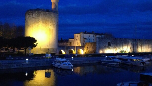 A historic tower with a prominent light at the top, situated next to a body of water with several boats docked.