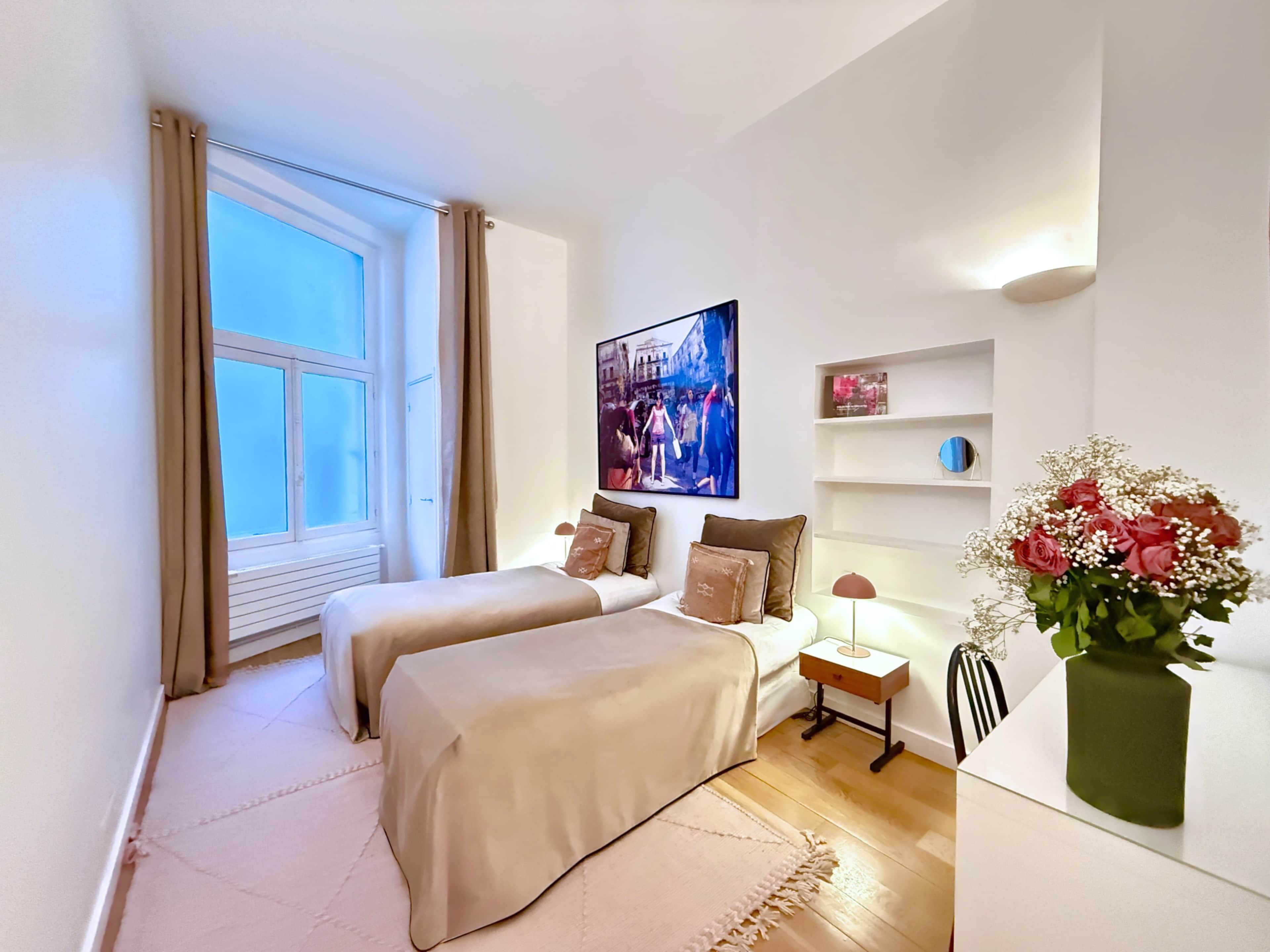 Una camera da letto con due letti, una finestra, un quadro, un comodino, una lampada e un vaso con fiori.