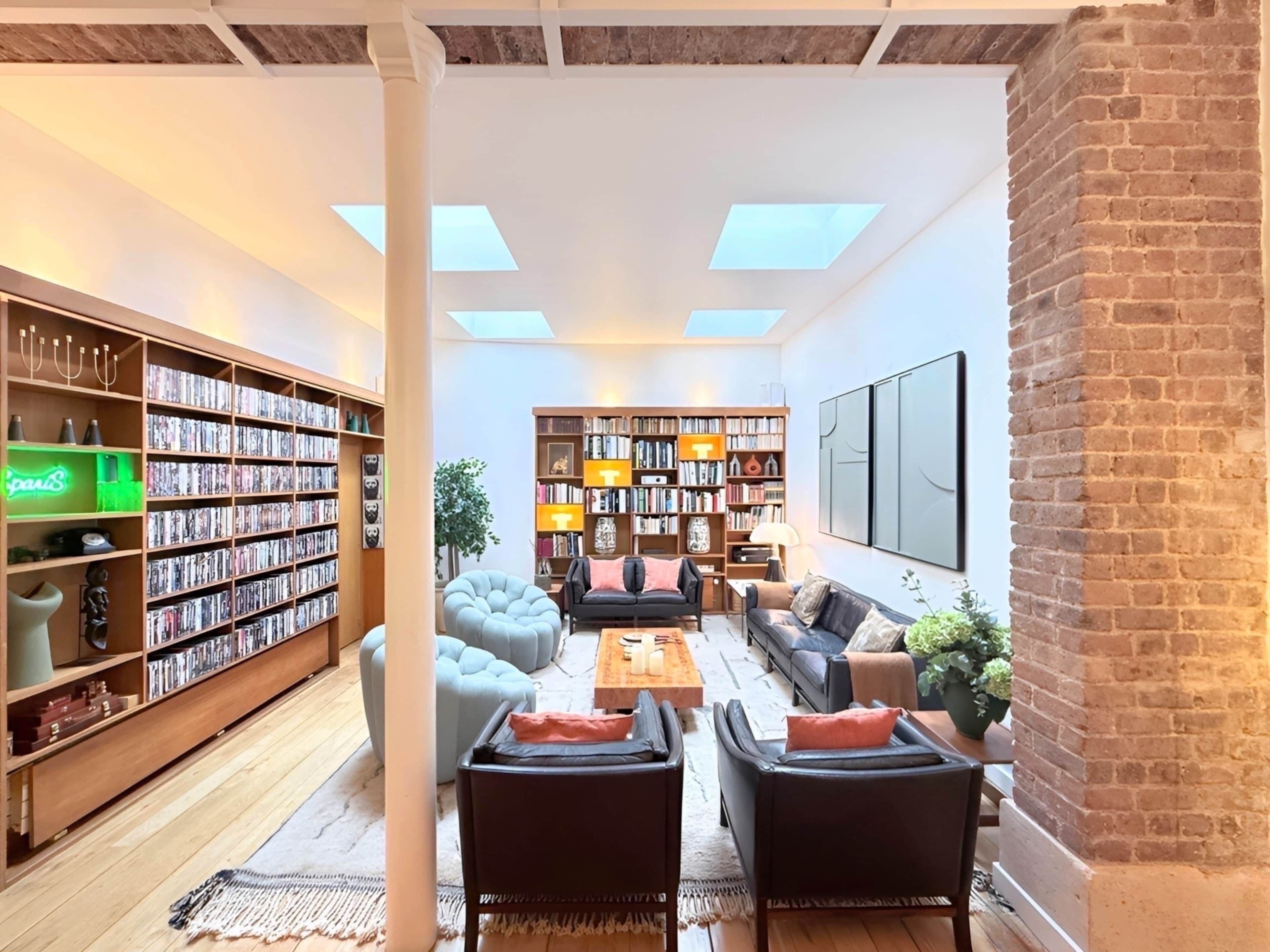 A spacious living room with a high ceiling, large windows, and a brick wall. There are two large bookshelves filled with books and decorative items, a comfortable seating area with sofas and a coffee table, and a large flat-screen TV mounted on the wall.