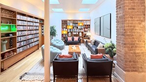 A spacious living room with a high ceiling, large windows, and a brick wall. There are two large bookshelves filled with books and decorative items, a comfortable seating area with sofas and a coffee table, and a large flat-screen TV mounted on the wall. - Sparissimo, 6 Br / 4 Ba, 14 ppl, 220 m² (Paris)