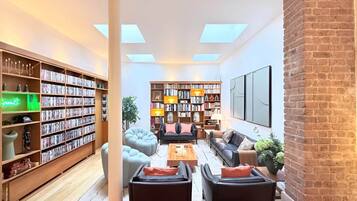 A spacious living room with a high ceiling, large windows, and a brick wall. There are two large bookshelves filled with books and decorative items, a comfortable seating area with sofas and a coffee table, and a large flat-screen TV mounted on the wall.