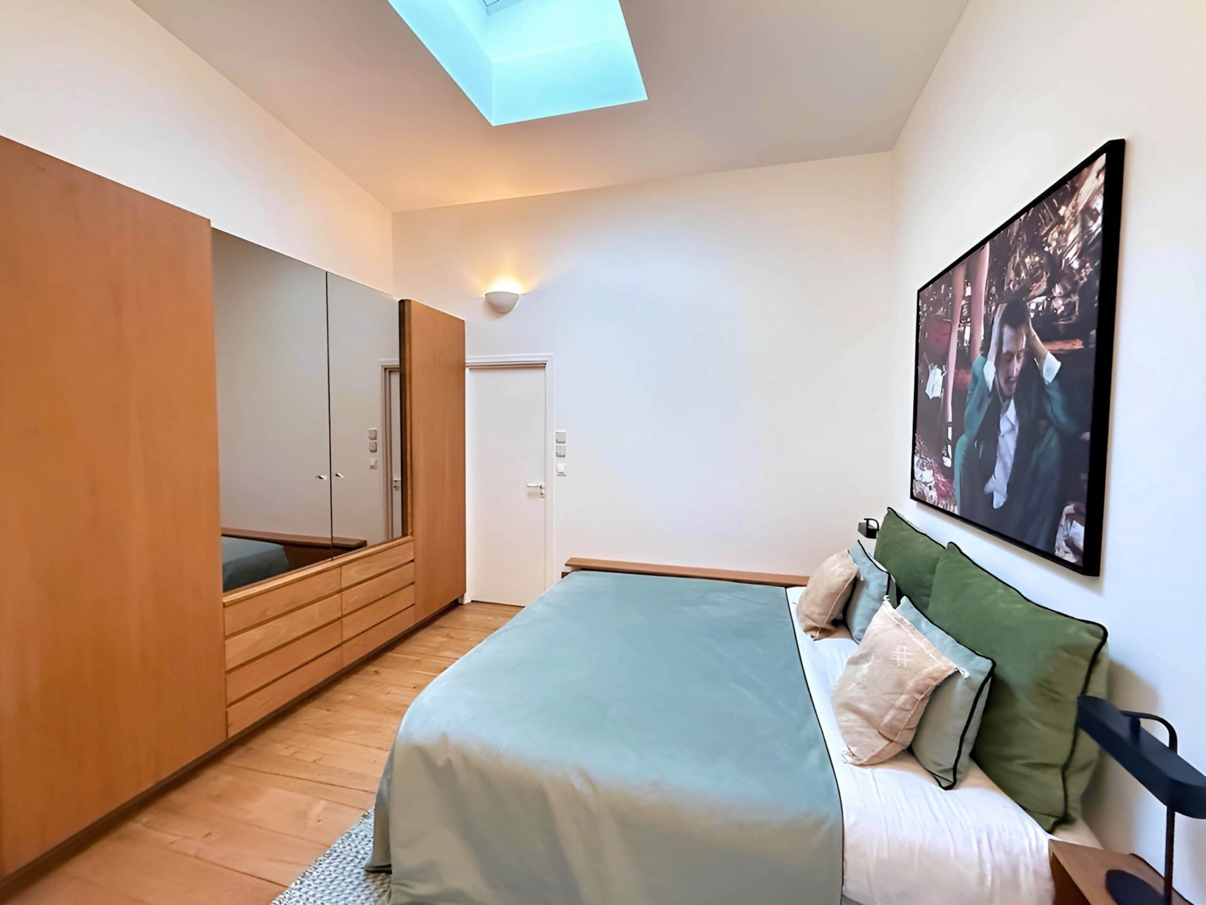 A bedroom with a bed, a wardrobe, a skylight, and a painting on the wall.