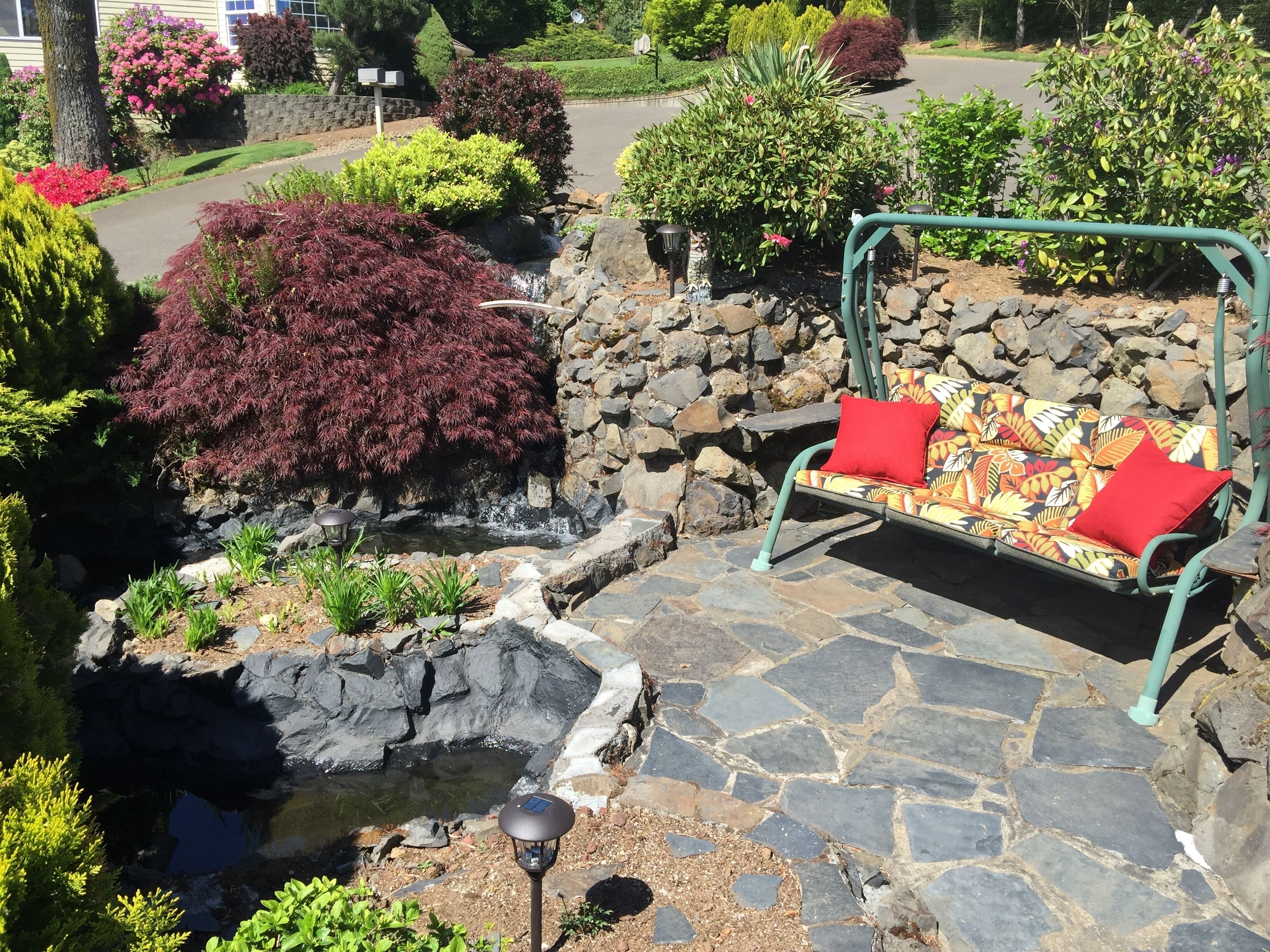Un jardí amb un camí de pedra, una petita bassa i una cadira amb balancí amb coixins vermells.