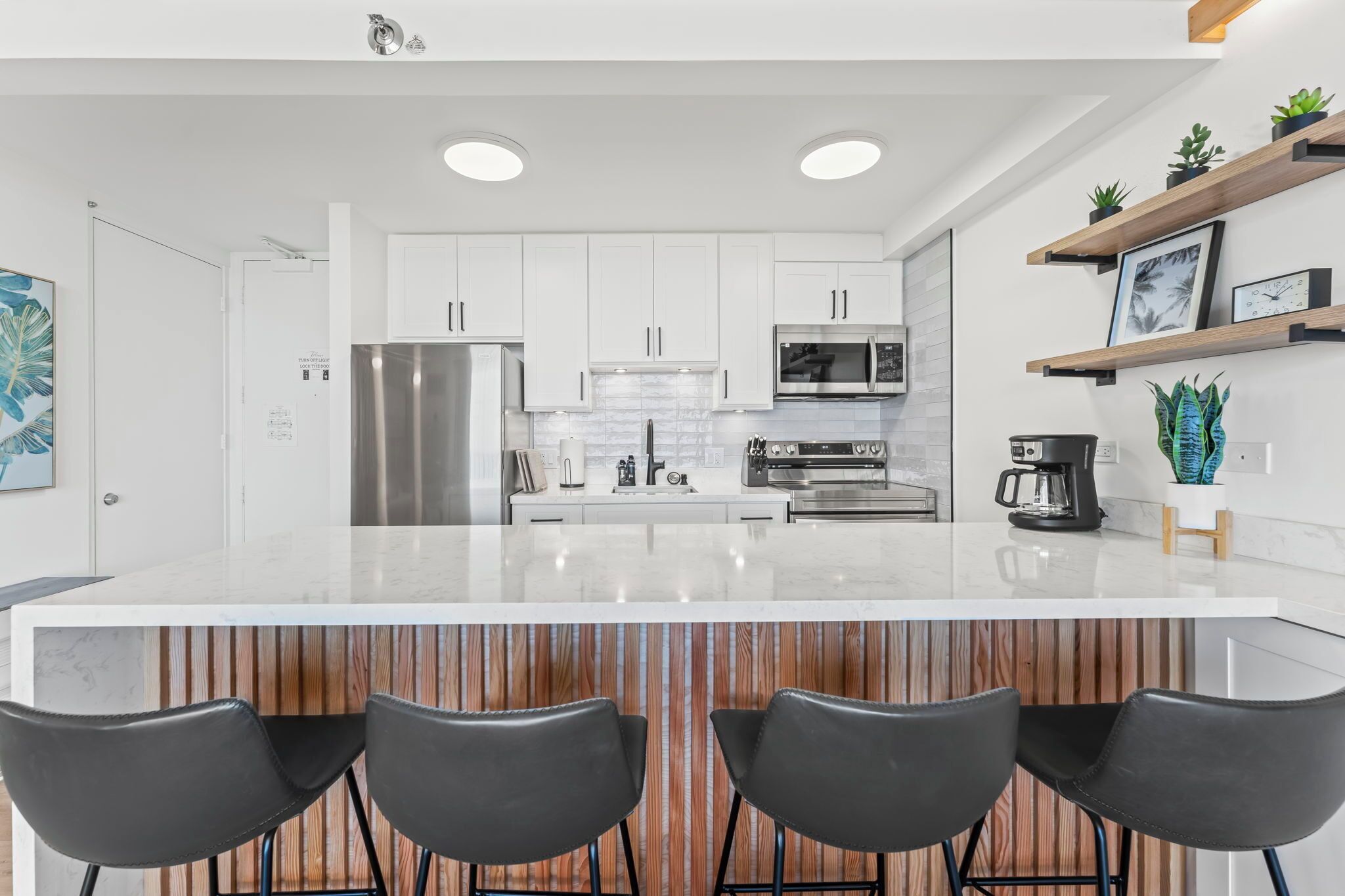 Una cocina moderna con armarios blancos, electrodomésticos de acero inoxidable, una isla grande con taburetes y plantas decorativas.