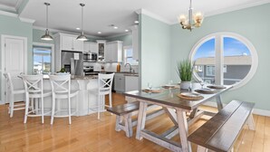 A spacious kitchen with white cabinetry, a central island, and a dining area with a wooden table and benches. - " 'Til Next Time!" Pool w/Opt. Heat, Hot Tub, Tiki Bar, Elevator, Crow's Nest (Holden Beach)