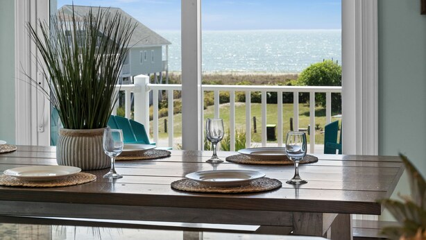 A dining table set with plates and glasses, overlooking a coastal view with a white fence and greenery. - " 'Til Next Time!" Pool w/Opt. Heat, Hot Tub, Tiki Bar, Elevator, Crow's Nest (Holden Beach)