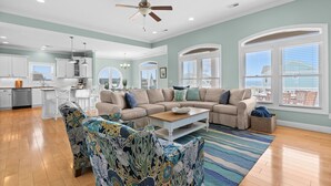 A spacious living room with a large sectional sofa, a patterned armchair, and a coffee table. There is a kitchen in the background with white cabinets and a dining area. - " 'Til Next Time!" Pool w/Opt. Heat, Hot Tub, Tiki Bar, Elevator, Crow's Nest (Holden Beach)