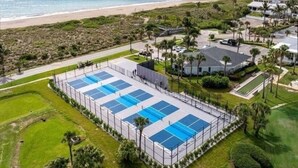 A large, rectangular swimming pool with multiple lanes, surrounded by a fence and palm trees, with a beach and ocean in the background. - Ocean Front Community- Ocean Village at South Hutchinson Island- Treasure Coast (Fort Pierce)