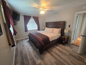 A bedroom with a wooden bed, a ceiling fan, a TV mounted on the wall, and a window with curtains.