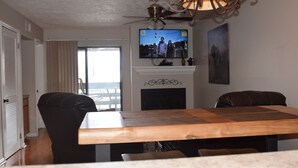 A dining area with a wooden table, black chairs, a fireplace, a television mounted above it, and a view of the outdoors through a large window.