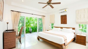 Un dormitorio amplio con una cama grande, un ventilador de techo, un equipo de aire acondicionado y vistas a la vegetación a través de las ventanas.
