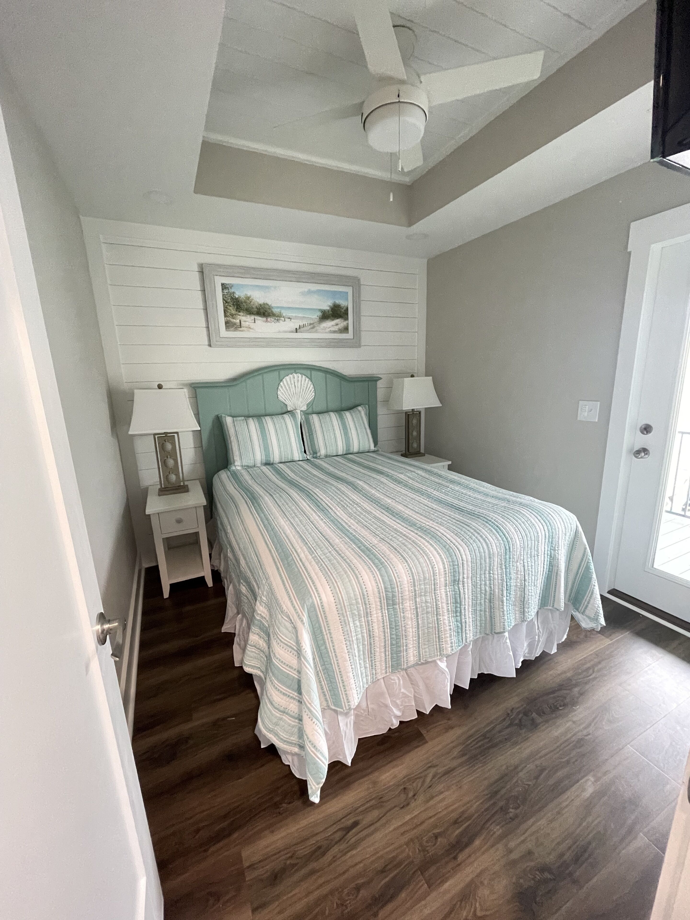 Une chambre à coucher comprenant un lit, deux tables de chevet, un ventilateur de plafond, un tableau encadré accroché au mur et une porte.