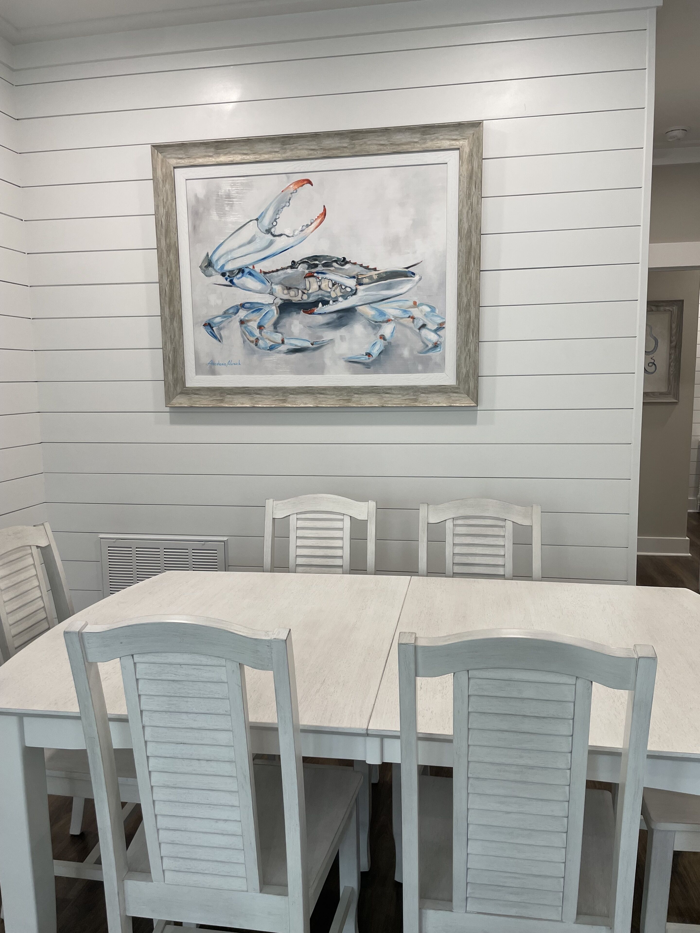 Un coin repas avec une table et des chaises blanches, un tableau encadré représentant un crabe accroché au mur, et un plafond blanc orné de lignes horizontales.