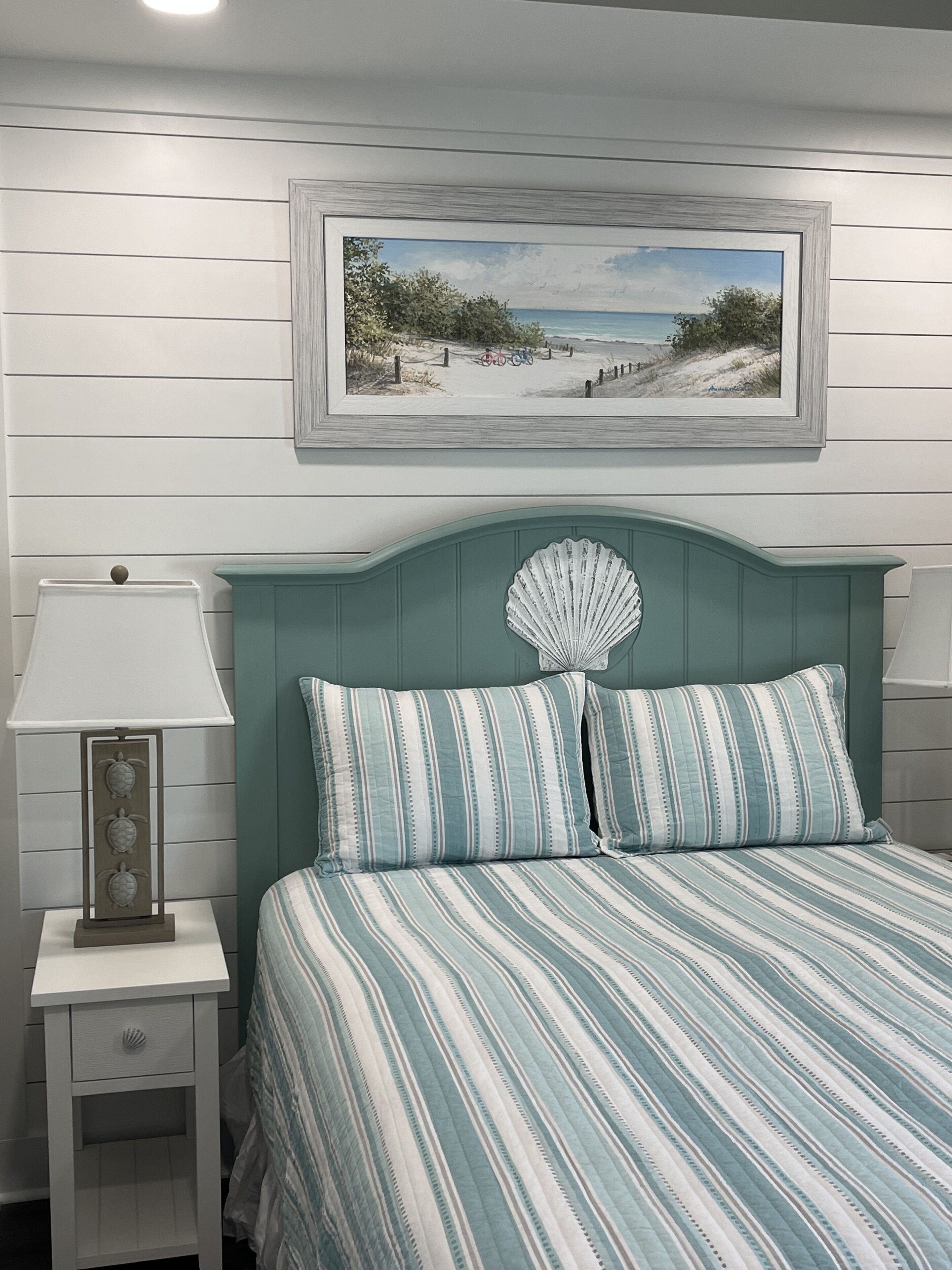 Ein Schlafzimmer mit gestreifter Bettdecke, einem gerahmten Strandbild und einer dekorativen Muschel am Kopfteil.