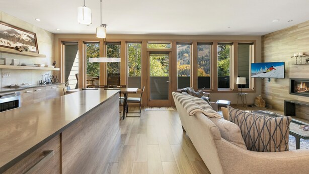 A modern kitchen with a large island, a dining area with a table and chairs, a living room with a sofa and a fireplace, and large windows offering a view of the outdoors.