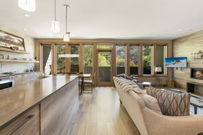 Una cocina moderna con una isla grande, un comedor con mesa y sillas, una sala de estar con sofá y chimenea, y ventanas amplias que ofrecen vistas al exterior.