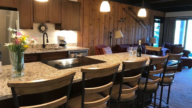 A kitchen with a granite countertop, wooden bar stools, a coffee maker, and a dining area with a table and chairs. - Remodeled ski in/ski out condo in Mt Crested Butte with incredible views! (Crested Butte)