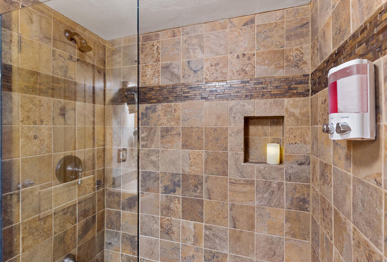 A shower with a glass door, a soap dispenser, and a candle in a niche.