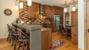 A modern kitchen with wooden cabinets, a central island with bar stools, a stainless steel refrigerator, and a patterned backsplash. - Fairway Nine Town Home Retreat Sun Valley (Sun Valley)