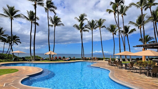 A swimming pool with a curved edge, surrounded by palm trees and lounge chairs with umbrellas. - Largest Two Bedroom/ Two Bath in the Village, Sleeps Eight & Close to the Beach (Wailea)