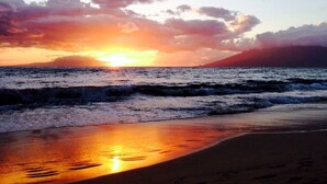 A beach with a sunset over the ocean, waves approaching the shore, and a mountain in the background. - Largest Two Bedroom/ Two Bath in the Village, Sleeps Eight & Close to the Beach (Wailea)