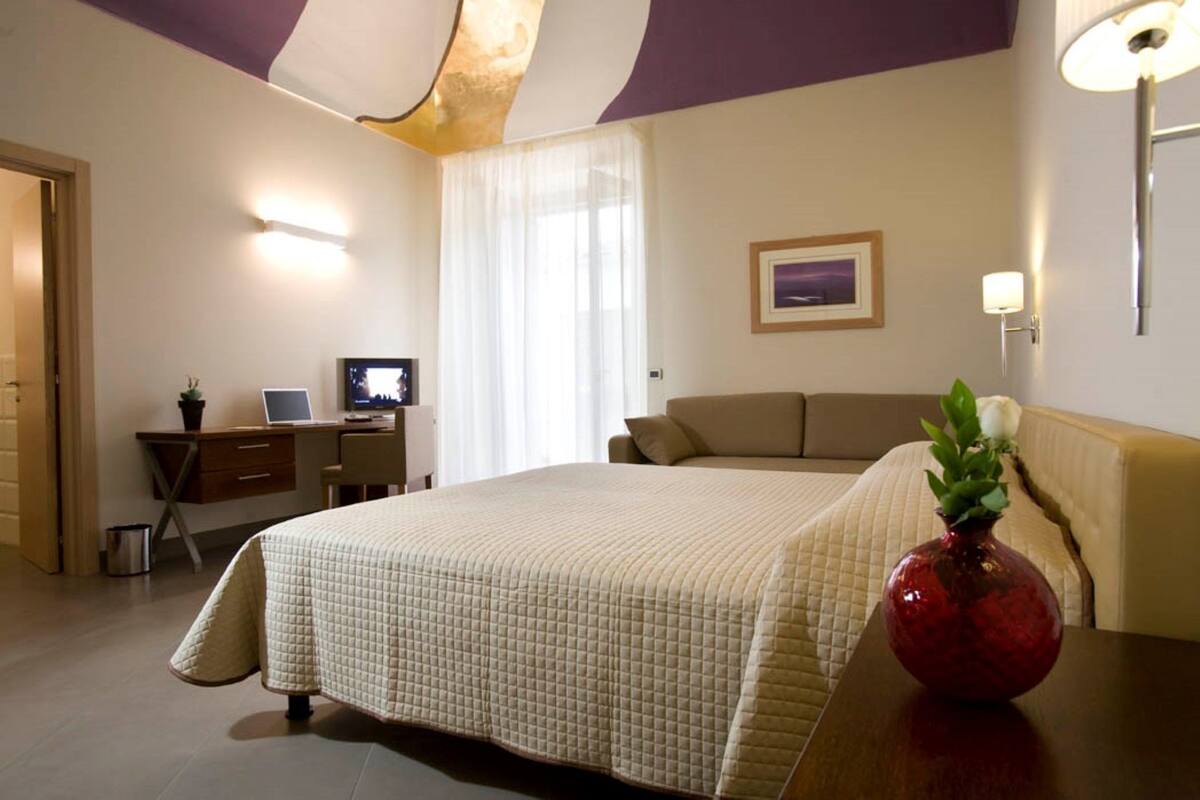 A hotel room with a large bed, a desk with a computer, a small potted plant, and a vase on the nightstand.