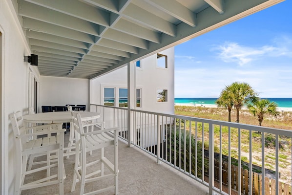 A balcony with a view of the beach and ocean, white furniture, and a white building with windows.