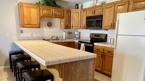 A kitchen with wooden cabinets, a white refrigerator, a microwave, and a stove. There are bar stools and a large island with a sink. - Las Palmas Spectacular View-Elevator, Washer-Dryer, No Resort Fees - Great RATE! (St George)